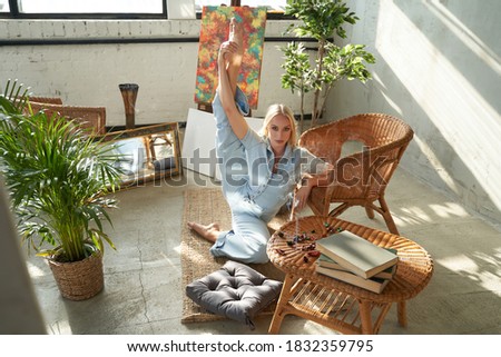 Beautiful and flexible woman with blond hairs in styled clothes posing on carpet with raised foot around bamboo furniture in living room.