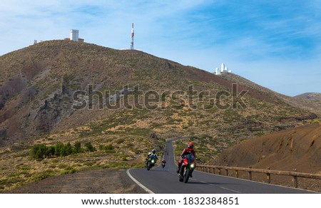 A winding mountain road in the Teide region. A group of motorcyclists sped across the asphalt. In the background the Tenerife observatory on the mountain with blue sky and clouds.