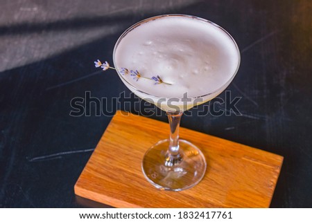 Summer cocktail in a Martini glass with fresh lavender flower. Lavander alcohol drink, refreshing beverage in restaurant. On black wooden table over blurred background.