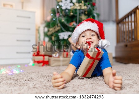 happy child boy on Christmas morning recieve present, laying on cozy carpet in his room in pajama and red santa hat, open gifts. New Year tree on background