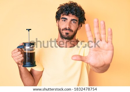 Handsome young man with curly hair and beard holding french coffee maker with open hand doing stop sign with serious and confident expression, defense gesture 