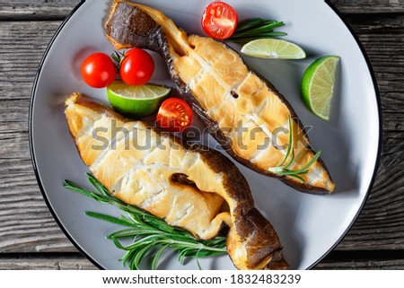 Traditional wild-caught smoked halibut fish steaks with lime, rosemary sprigs and tomatoes on a plate on a dark wooden background, top view, close-up