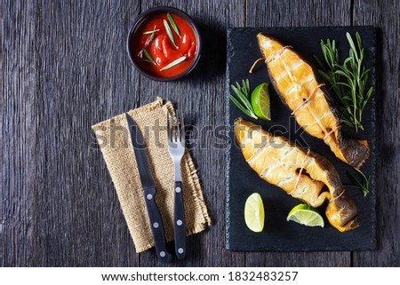 Traditional smoked halibut fish steaks with lime and rosemary sprigs on a black slate on a dark wooden background, top view, close-up