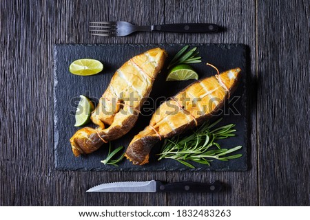 Traditional smoked halibut fish steaks with lime and rosemary sprigs on a black slate on a dark wooden background, top view, close-up
