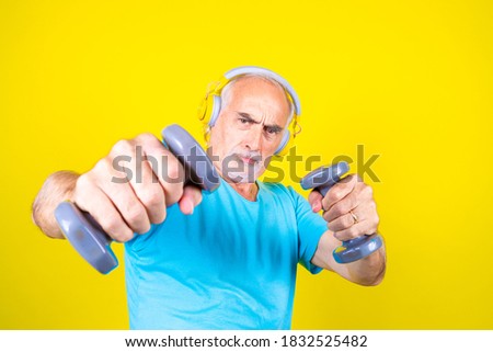 Elderly man training using dumbbells on yellow background determined - Isolated mature man stretching using gym weight listening music - healthy, sport, training concept