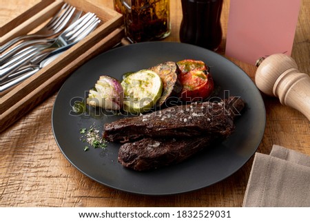 Roasted beef fillet, dark and yummy, with grilled vegetables as a side dish, knife, fork and pepper shaker aside on a wooden table