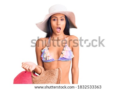 Young beautiful latin girl wearing bikini and hat holding bag scared and amazed with open mouth for surprise, disbelief face 