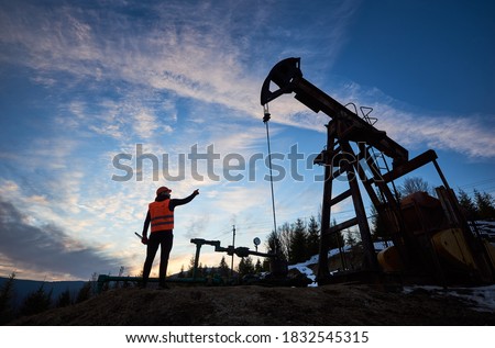 Back view of oil man in work vest standing near petroleum pump jack with sunset on background. Oil worker pointing finger at oil pump rocker-machine. Concept of oil extraction and petroleum industry.