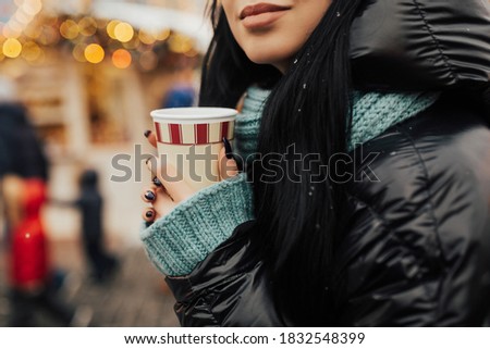 Girl in black jacket and a knitted scarf is holding paper glass in hands. She drinks a warm drink, coffee or tea at the Christmas market. In the background, lights flicker. Close up, cropped photo.
