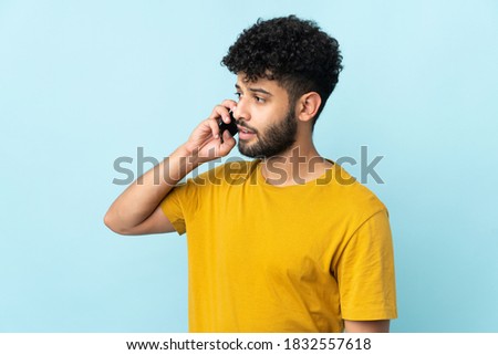 Young Moroccan man isolated on blue background keeping a conversation with the mobile phone with someone