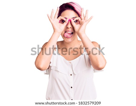 Young beautiful woman with pink hair wearing casual clothes and glasses doing ok gesture like binoculars sticking tongue out, eyes looking through fingers. crazy expression. 