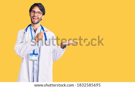 Handsome hispanic man wearing doctor uniform and stethoscope amazed and smiling to the camera while presenting with hand and pointing with finger. 