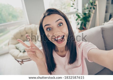 Photo portrait of young girl showing v-sign taking selfie sitting on sofa with open mouth indoors