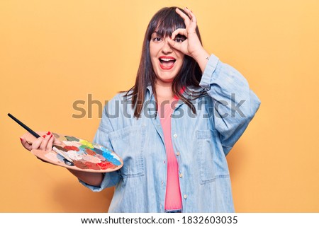Young plus size woman holding paintbrush and palette smiling happy doing ok sign with hand on eye looking through fingers 