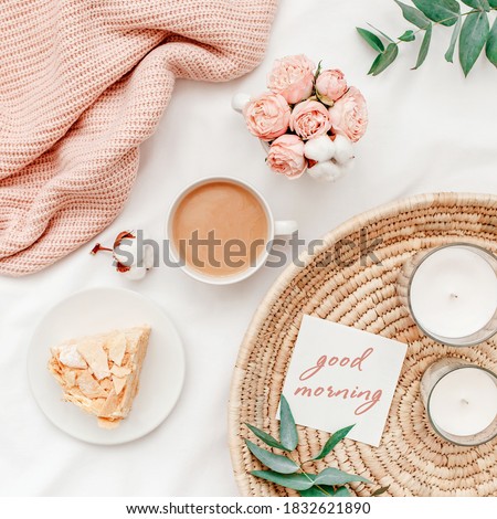 Wicker tray, cup of coffee, piece of cake, rose flowers, eucalyptus branch, candles, pink knitted plaid or blanket, card with text GOOD MORNING. Breakfast in bed. Stylish home interior decor. Top view