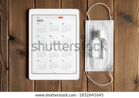 a tablet with an open calendar for 2021 year and protective medical mask and hand sanitizer on a wooden boards table background. covid-19 coronavirus protection concept in 2021 Royalty-Free Stock Photo #1832641645