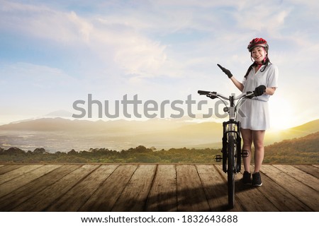 An Asian woman with a bicycle helmet standing beside her bicycle on a wooden path with a landscape background