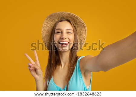 Cheerful young woman in summer hat tourist taking selfie on yellow studio background, showing peace gesture. Happy travel blogger taking photo of herself while travelling around the world