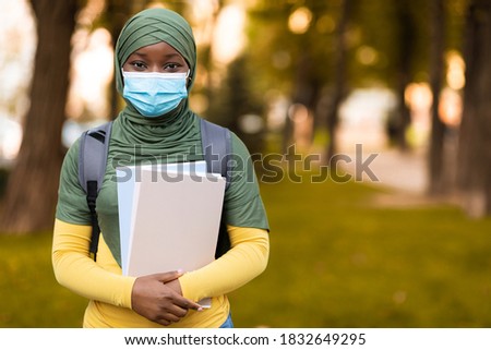 Covid-19 Education. Black muslim female student wearing medical mask and hijab posing outdoors, holding backpack and books, standing on autumn street, looking at camera, selective focus, copy space