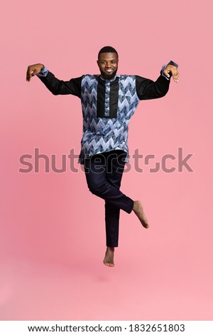 Carefree black man in african costume posing while jumping up on pink studio background. Smiling african american guy wearing traditional clothes, full size photo. Traditions and culture concept