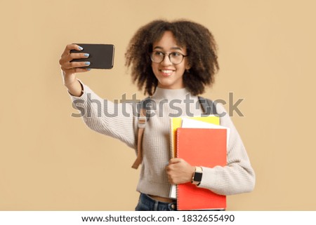 Photo for educational blog. Smiling smart african american young lady in glasses with backpack and smart watch holding notebooks and makes selfie isolated on yellow background, studio shot, copy space