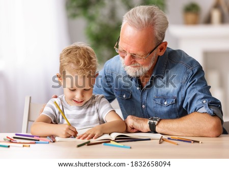 Happy boy smiling and looking at bearded elderly man while sitting at table and drawing picture in sketchbook
