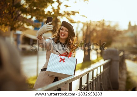 Hello autumn. smiling elegant woman in beige trench coat and black beret with shopping bags and autumn yellow leaves outdoors in the city in autumn taking selfie with cellphone.