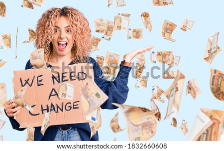 Young blonde woman with curly hair holding we need a change banner celebrating victory with happy smile and winner expression with raised hands