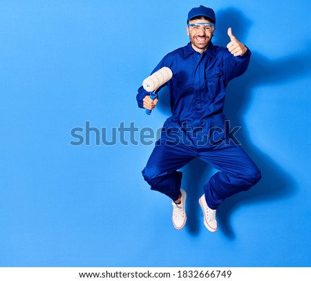 Young handsome hispanic man wearing painter uniform and glasses smiling happy. Jumping with smile on face holding roller doing ok sign with thumb up over isolated blue background