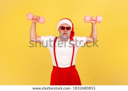 Assertive funny man in santa claus costume rising hands holding dumbbells, pumping up muscles, holiday discounts at gym. Indoor studio shot isolated on yellow background