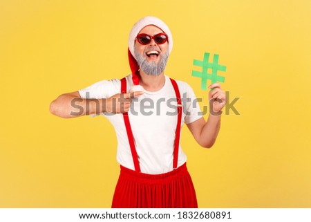 Happy smiling gray bearded man in sunglasses and santa claus hat pointing at green hashtag symbol in his hand, tagging holiday posts. Indoor studio shot isolated on yellow background