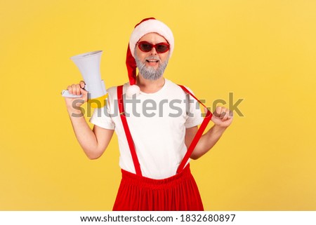 Positive self confident adult man in santa claus costume holding megaphone and looking at camera, ready to organize your holiday party. Indoor studio shot isolated on yellow background