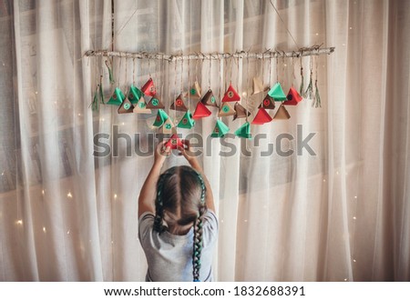Cute little kid opening handmade advent calendar with color paper triangles. Sweets and candies are hidden in colorful triangles hanging on branch. Seasonal activity for kids, family winter holidays