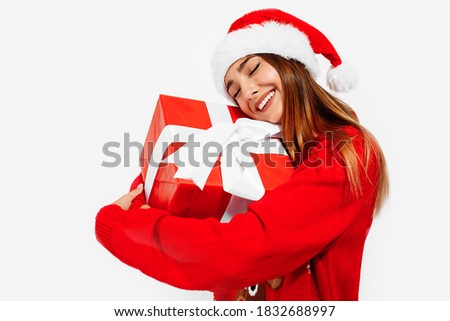 Happy cute young woman in christmas sweater and santa claus hat hugging present isolated on white background