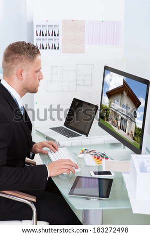 Businessman or estate agent checking a property portfolio online while sitting at his desk in the office looking at the exterior of a rural house visible on the desktop monitor