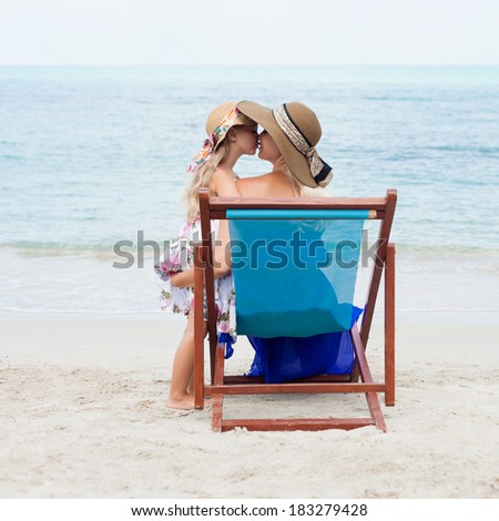 Beautiful little girl kissing her young mother sitting in a chair on the beach. Outdoors