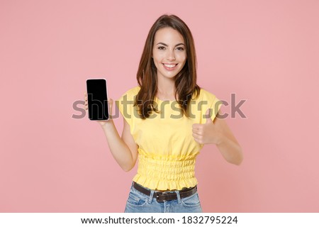 Smiling young brunette woman 20s wearing yellow casual t-shirt hold mobile cell phone with blank empty screen mock up copy space showing thumb up isolated on pastel pink background studio portrait
