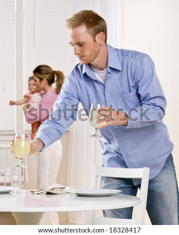 Young father setting table as mother holds baby daughter in dining room