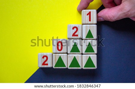 Wood cubes with number '2021' and Christmas tree icons stacking as step stair on beautiful yellow and black background, copy space. Male hand. Business concept.