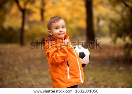 Little football player: toddler boy in orange jacket standing half turned and holding soccer ball in autumn park or forest. Smiling child with ball outdoors. Yellow autumn trees in background