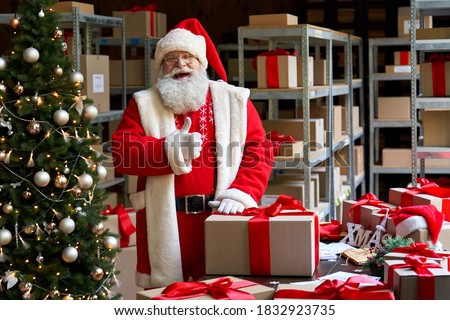 Happy old funny Santa Claus wearing costume holding Merry Christmas present gift box looking at camera showing thumbs up like standing in workshop warehouse. Fast xmas delivery concept. Portrait Royalty-Free Stock Photo #1832923735