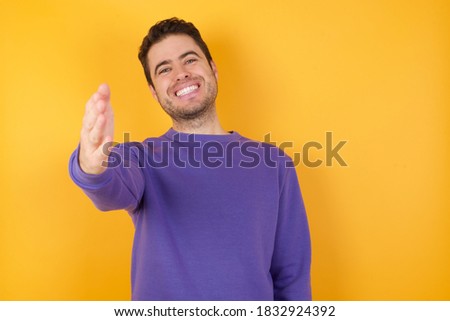 Handsome man with sweatshirt over isolated yellow background smiling friendly offering handshake as greeting and welcoming. Successful business.