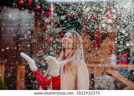 Young pretty blond woman in red Christmas sweater, beige winter coat and white mittens catching a snow outside in back yard with Christmas background.