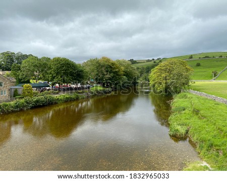 View looking down the River Wharfe, with old trees, cars, fields, and distant hills in, Burnsall, Skipton, UK