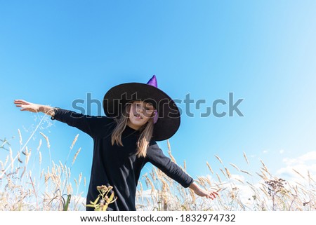 Girl in a witch costume flies with her arms outstretched to the sides. Halloween holiday. A child in a pointed hat. Dry grass field.