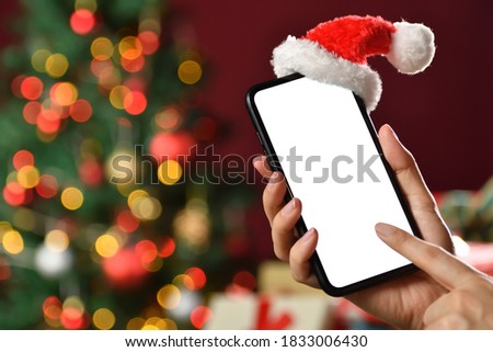 Hand holding the black smartphone with blank screen and Santa Claus' hat on top on Christmas background