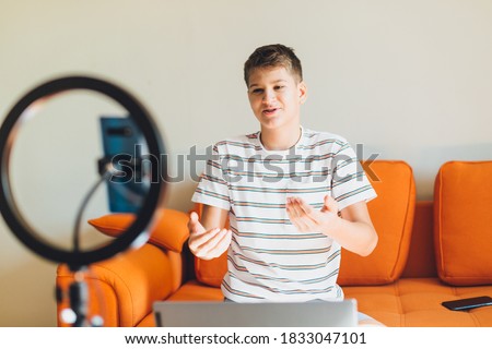 Young teenager blogger recording video at home. Cheerful cute boy pointing with hands into smartphone. Boy with camera recording video on orange couch.