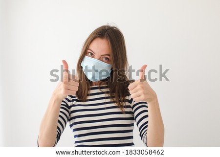 Adult female in medical mask looking at camera and showing thumb up gesture against gray wall during epidemic