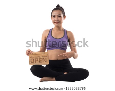 Pretty Asian woman in exercising clothing sitting with one hand pointing on the wooden plate saying Welcome. We're OPEN on white background. Class opening again.