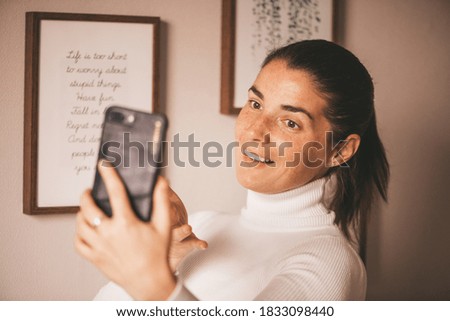 Pretty young smiled Caucasian woman in white turtleneck smiling and videochatting on mobile phone in home. Stylish female having video call on smartphone. Girl talking. Videochat in social media.